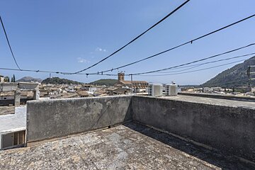 Rooftop view over a town with red-tiled roofs, a church, and mountains under a clear sky. Power lines crisscross, and AC units sit on the roof.