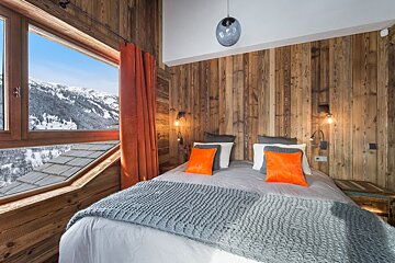 A cozy, rustic bedroom with wooden walls, orange accents, and a large window showcasing a stunning snowy mountain view.