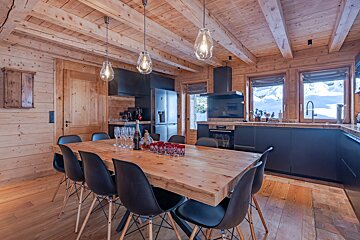 Rustic wooden chalet interior with modern black kitchen, large dining table, and snowy window views.
