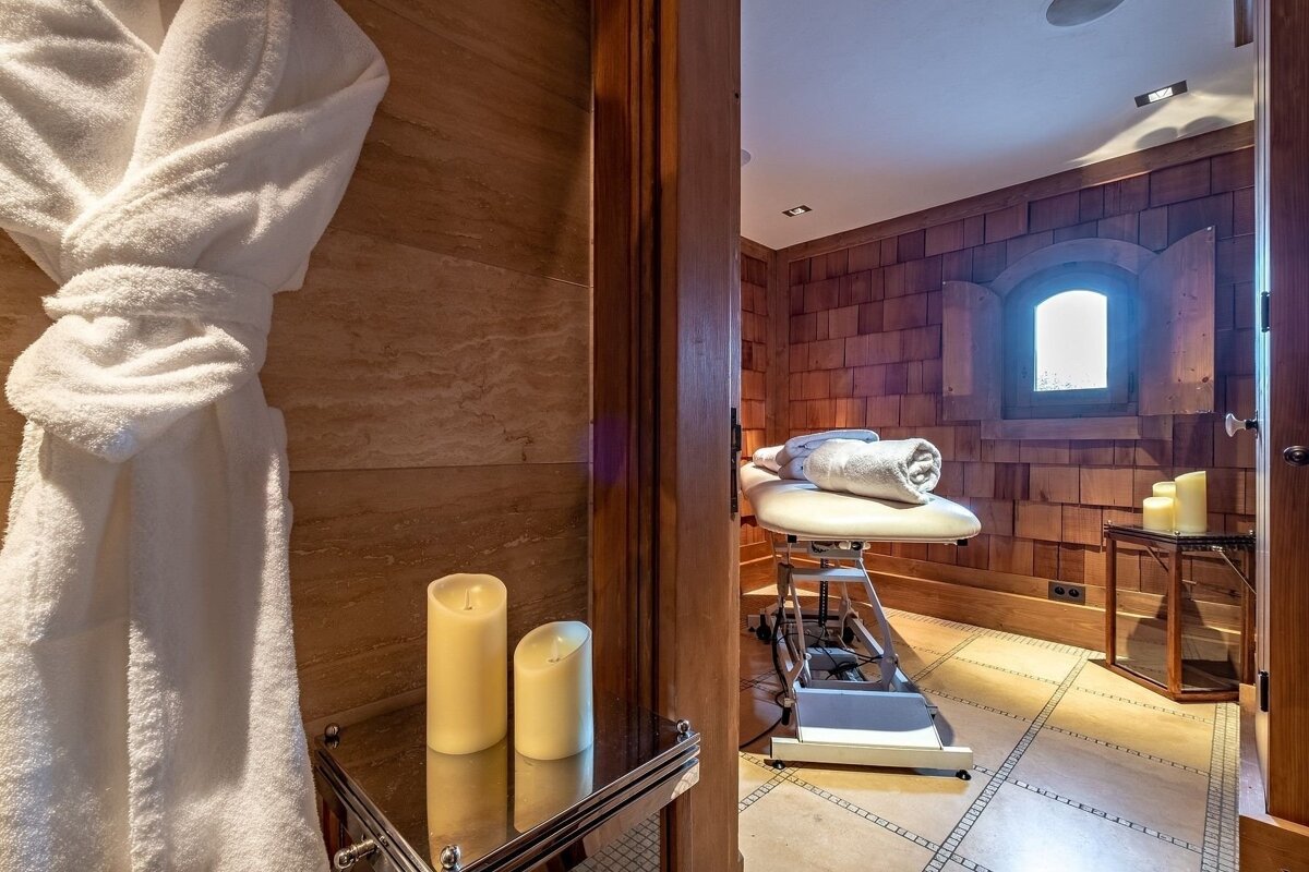 A cozy spa room features a massage table with rolled towels, glowing candles, and a white robe. Wood-paneled walls and a small window create a tranquil atmosphere.