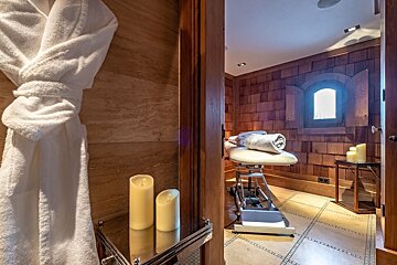 A cozy spa room features a massage table with rolled towels, glowing candles, and a white robe. Wood-paneled walls and a small window create a tranquil atmosphere.