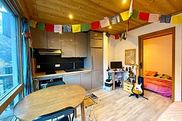 A cozy room featuring a grey kitchenette, a dining table, and a bedroom through a doorway. Prayer flags hang from the wooden ceiling.