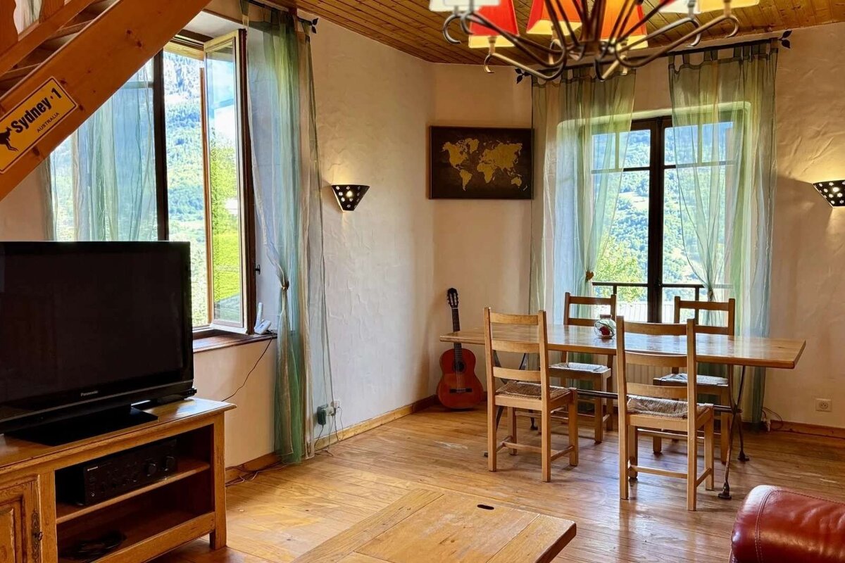 Cozy, rustic living-dining room with scenic mountain views through large windows. Features wooden furniture, TV, guitar, and a world map.