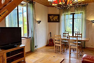 Cozy rustic living/dining room with large windows overlooking green mountains. Features a wooden table, TV, guitar, world map, and textured walls.