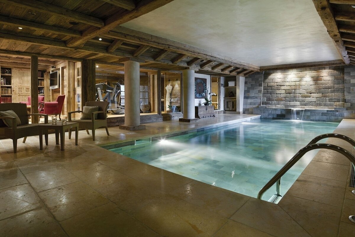 A luxurious indoor pool in a rustic chalet, featuring wooden beams, stone walls, a lounge, gym, and a waterfall feature.
