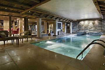 A luxurious indoor pool in a rustic chalet, featuring wooden beams, stone walls, a lounge, gym, and a waterfall feature.