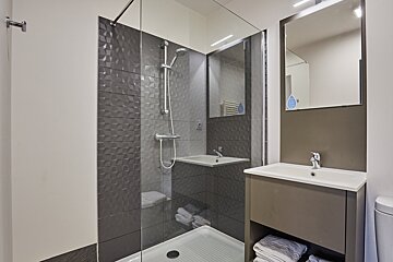 A modern bathroom features a glass shower with textured gray tiles, a sleek sink vanity, and a mirror. Towels are neatly stacked below the sink.