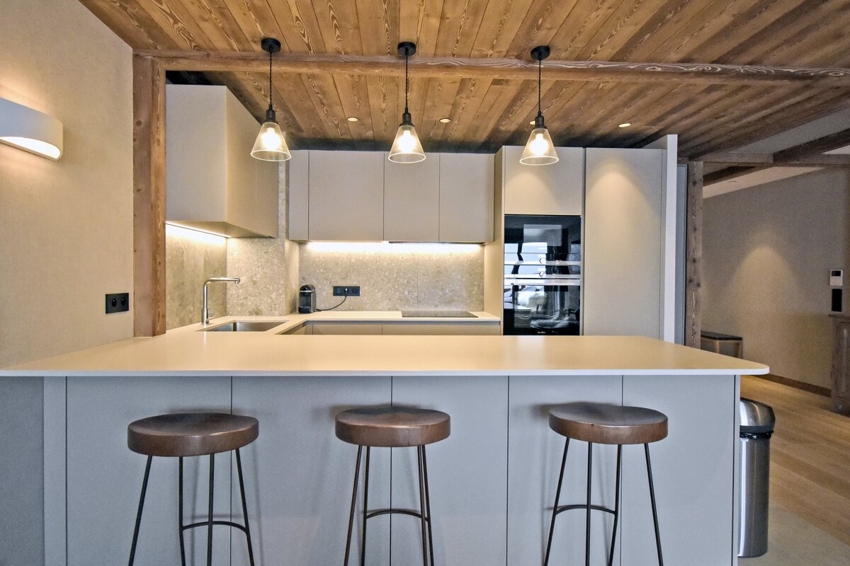 A modern kitchen features a large island with three bar stools, light grey cabinets, built-in appliances, and a warm wooden ceiling with pendant lights.