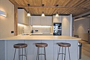 Modern minimalist kitchen featuring a beige island with three bar stools, hanging pendant lights, and a warm wood ceiling.