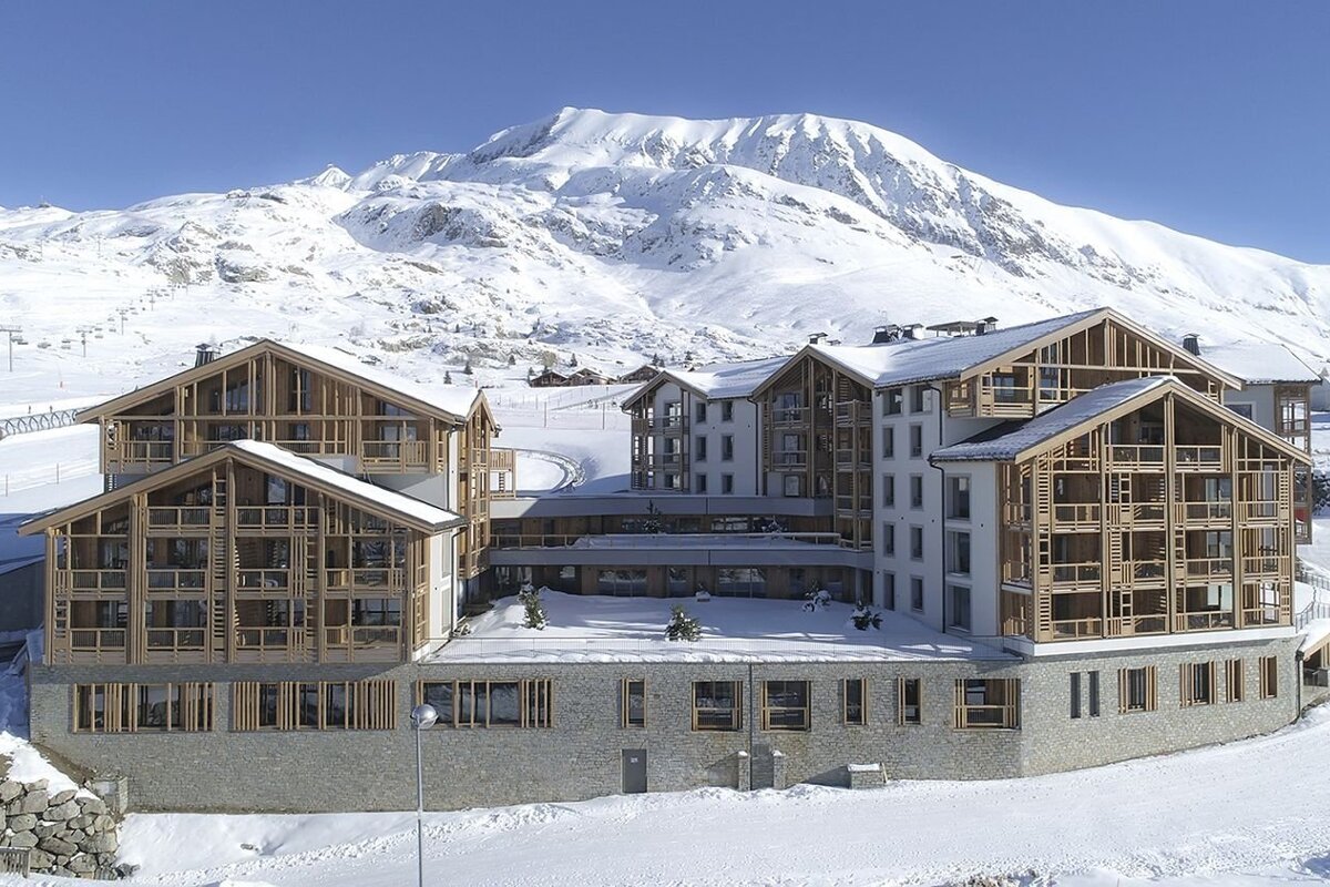 A large modern mountain resort with prominent wooden and stone architecture is surrounded by deep snow and towering, snow-capped peaks under a blue sky.