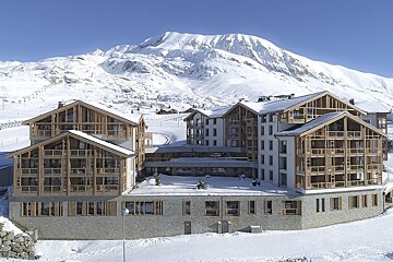A large modern mountain resort with prominent wooden and stone architecture is surrounded by deep snow and towering, snow-capped peaks under a blue sky.