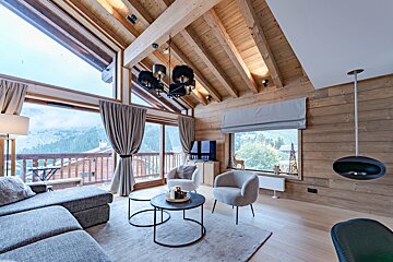 A warm, wood-paneled chalet living room featuring modern furniture, a vaulted ceiling, and stunning mountain views through large windows.