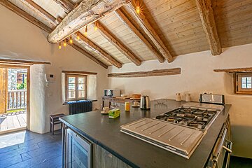 A rustic kitchen with exposed wooden ceiling beams, a gas stove, dark countertops, and an open doorway leading to an outdoor stone patio.