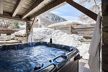 An outdoor hot tub bubbles under a wooden deck shelter, surrounded by deep snow with a bottle of champagne on the edge.
