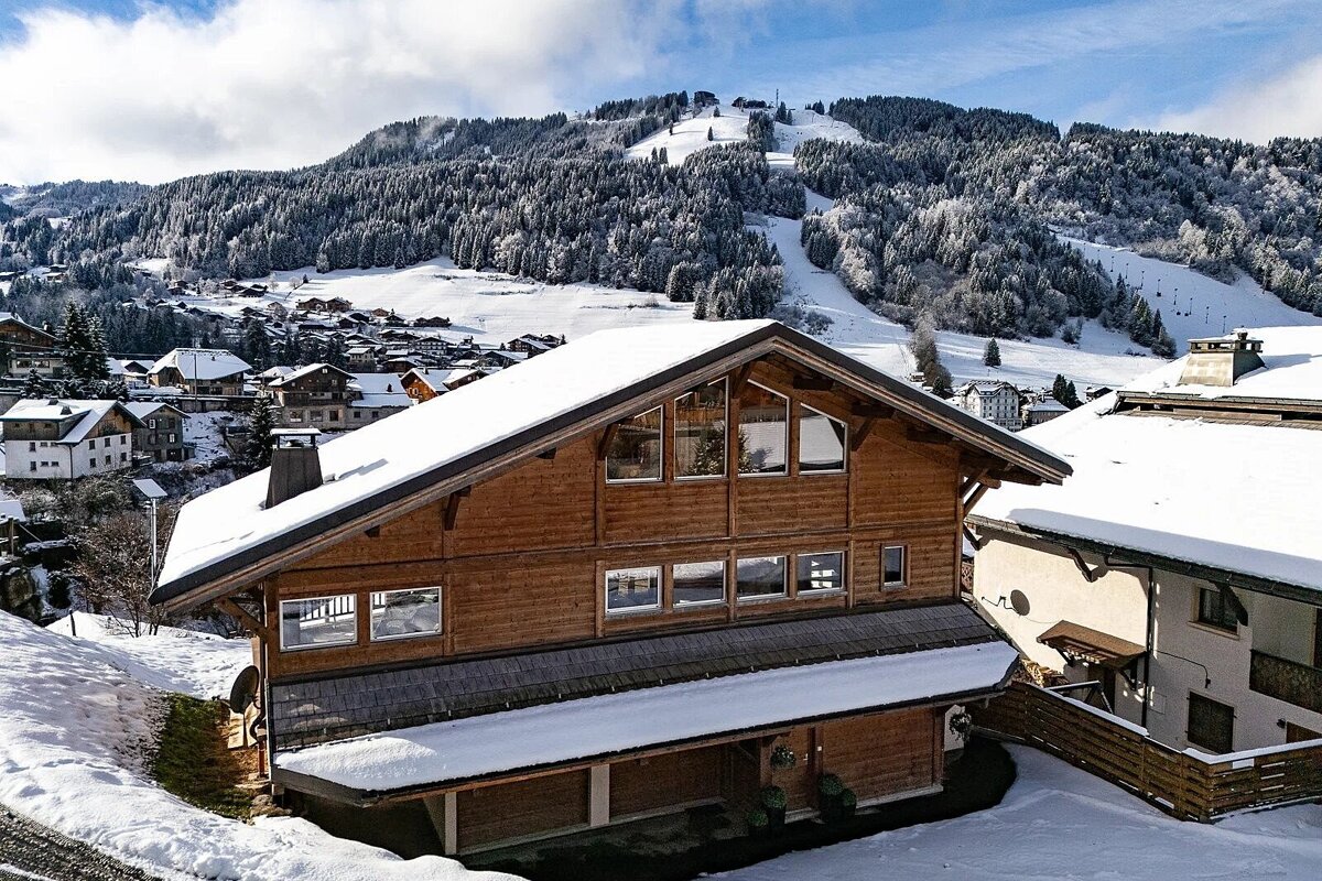 A large wooden chalet blanketed in snow, nestled in a mountain village with distant ski slopes visible under a bright sky.
