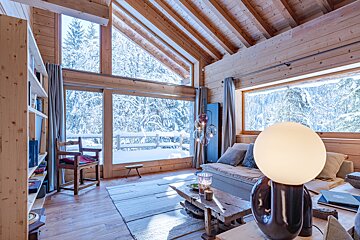 Cozy wooden cabin interior with large windows overlooking a snowy forest, featuring rustic furniture and a glowing globe lamp.