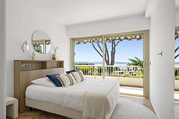 Serene bedroom with white decor, a chic wooden headboard, and expansive glass doors opening to a balcony with stunning ocean and tree views.