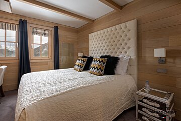 A cozy bedroom with wood-paneled walls, a large bed featuring a tufted headboard and patterned pillows. Windows frame a mountain view.