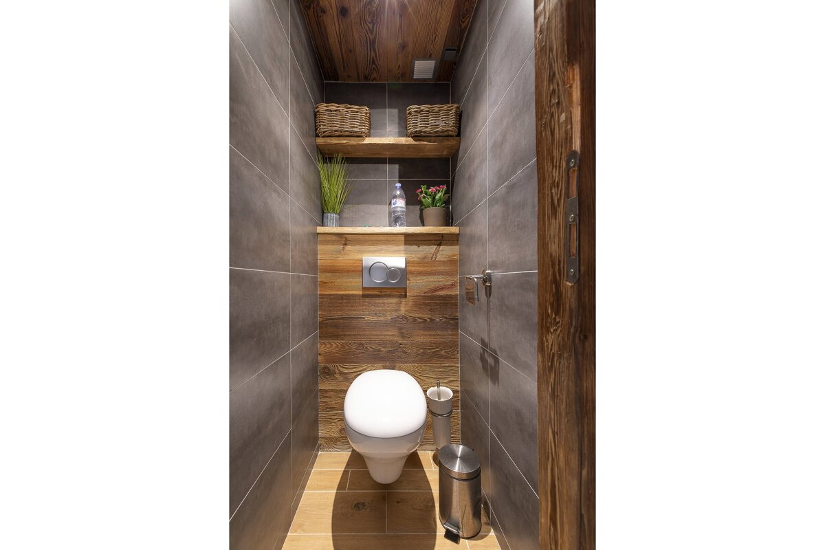 A stylish, narrow toilet room with grey tiled walls, warm wooden accents on the back wall, ceiling, and floor, featuring shelves with decor and a white toilet.