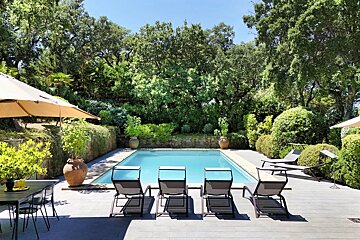 Serene outdoor swimming pool with sun loungers, a shaded dining area, and lush green landscaping under a bright sky.