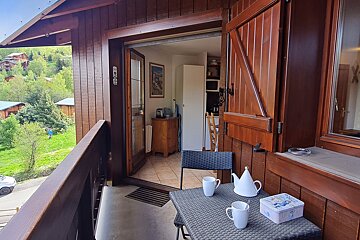 A cozy wooden balcony with a table, teapot, and mugs overlooking a green valley with houses. An open door reveals a kitchen area inside.