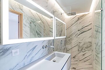 A sleek, modern bathroom featuring white marble walls, an illuminated mirror, a minimalist vanity, and a glass-enclosed shower.