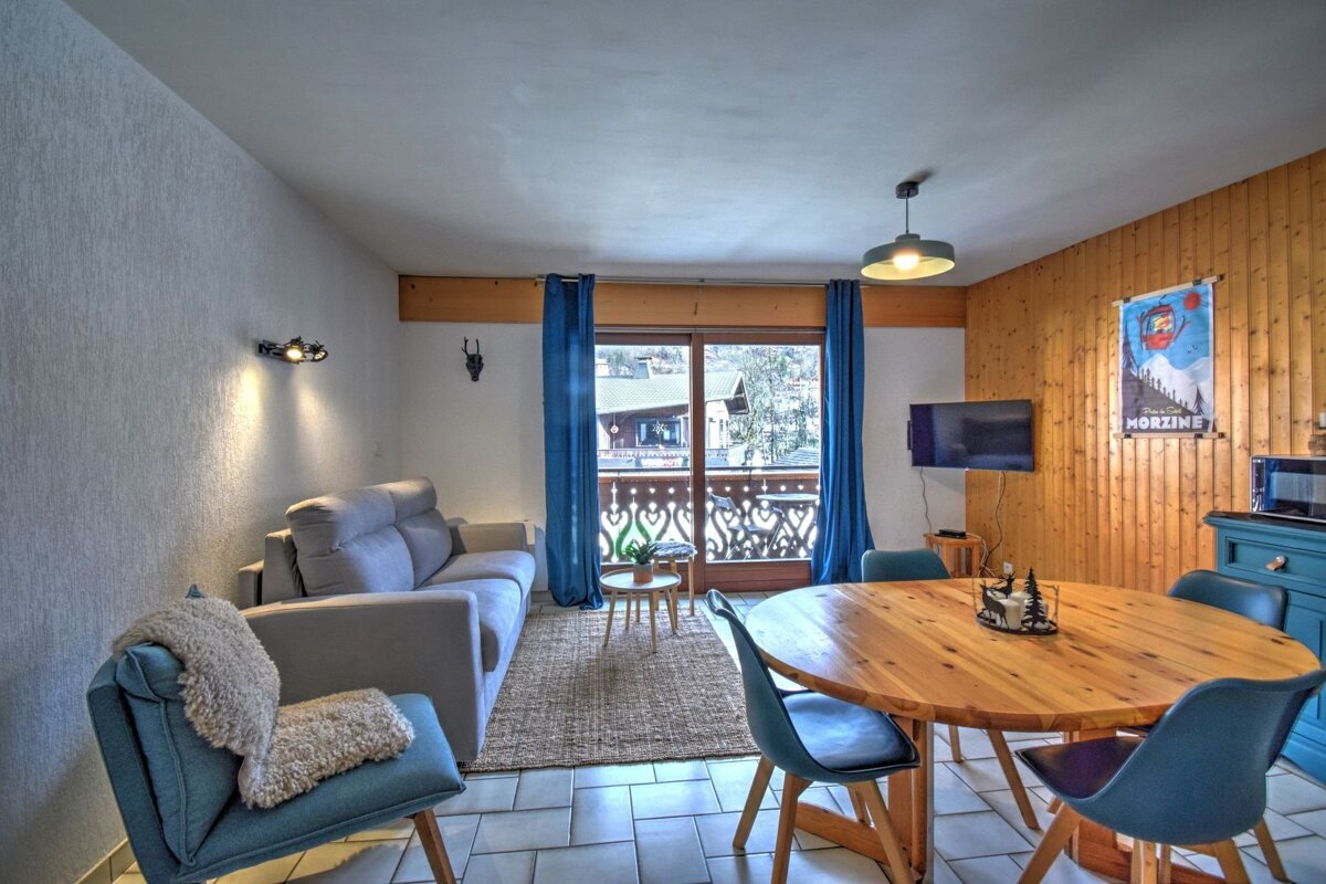 A cozy chalet-style living/dining room with a gray sofa, wooden dining table, blue chairs, and a balcony view. Features wood paneling and ski decor.