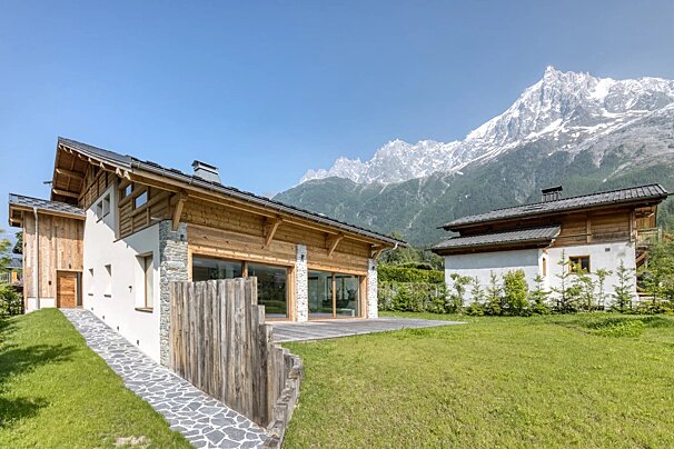 Modern chalets on a green lawn against a backdrop of towering, snow-capped mountains under a clear blue sky.