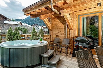 Cozy wooden deck of an alpine chalet featuring a hot tub, comfortable chairs, a BBQ grill, and scenic mountain views.