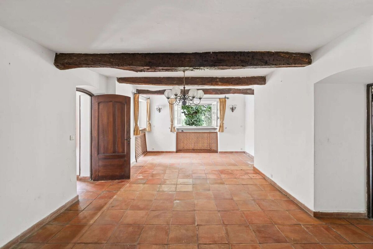 An empty room with white walls, terracotta floor, exposed wooden ceiling beams, a chandelier, and a window.