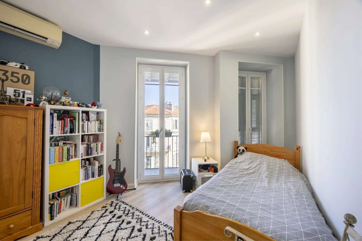 A bright bedroom with a single wooden bed, a white bookcase with yellow accents, and a red electric guitar. A balcony door and an air conditioner are visible.