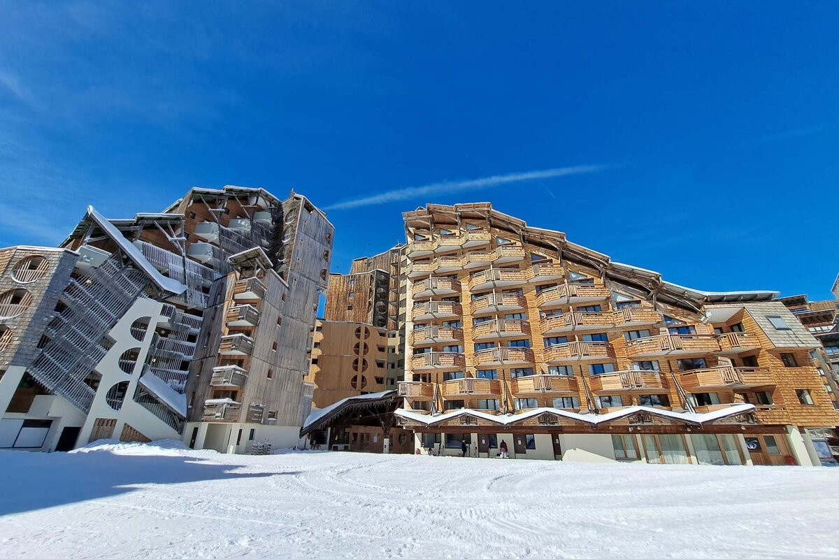Unique, modern wooden ski resort buildings rise from a snowy landscape under a clear blue sky, showing distinctive balconies and angles.