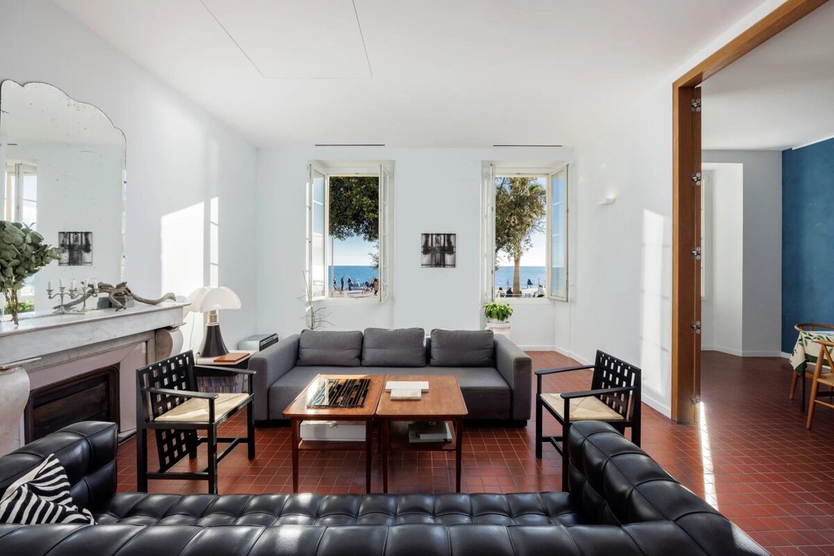 A sunlit living room with an incredible ocean view through two open windows. Features a gray sofa, black leather seating, and terracotta tile floors.