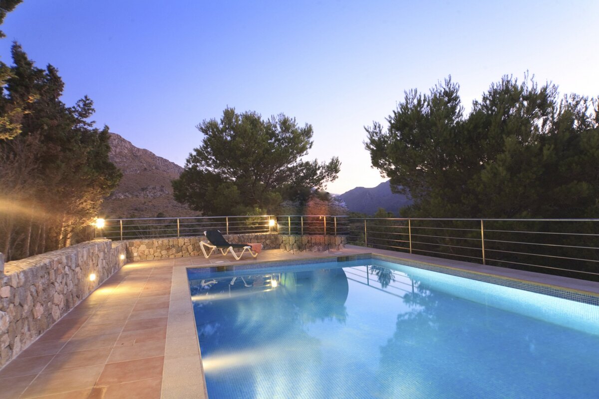 A beautifully lit swimming pool and patio at dusk, offering a serene mountain view. Pine trees frame the tranquil scene.