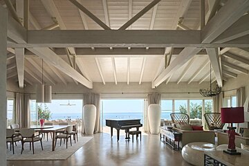 Spacious open-plan living room with high beamed ceilings, a grand piano, and large windows looking out to the ocean.