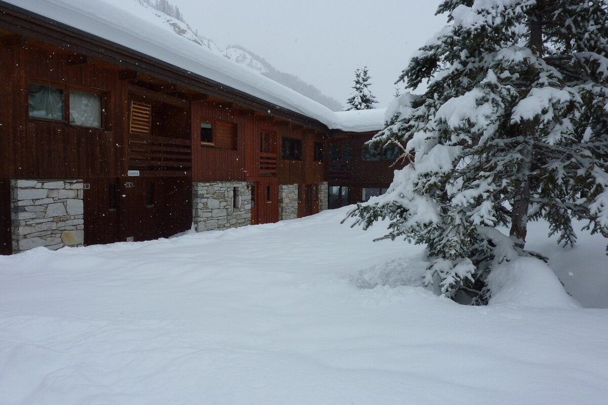 A snow-covered wooden chalet with stone bases and snow-laden pine trees in a wintry landscape with falling snowflakes.