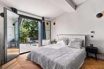 A modern, light-filled bedroom with a white bed, herringbone wood floors, and open glass doors leading to an outdoor patio surrounded by green trees.