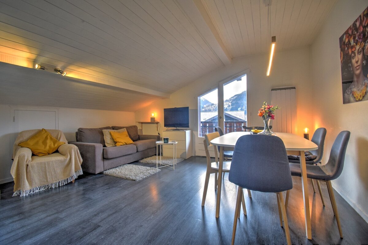 A cozy, modern attic apartment featuring a living and dining area, warm lighting, dark floors, and a window offering a scenic mountain view.