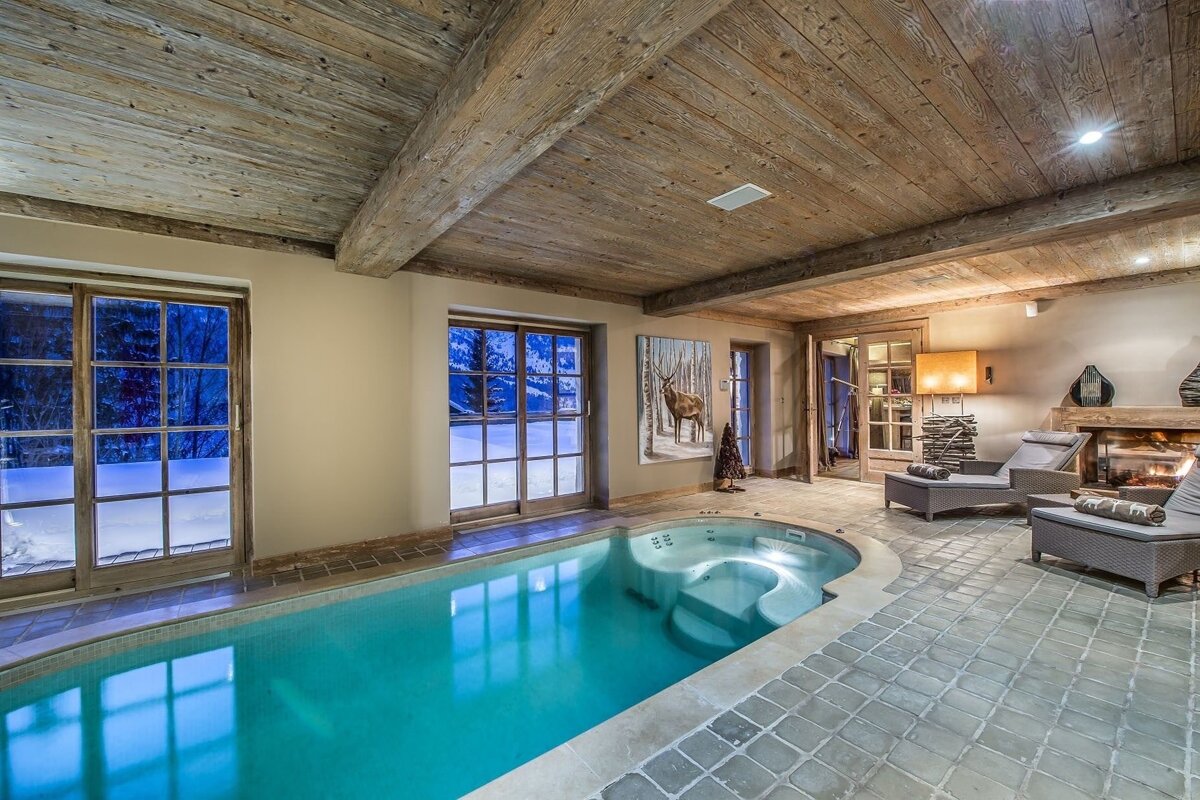 A luxurious indoor pool and jacuzzi room with rustic wood beams, stone floor, large windows to a snowy landscape, and a warm fireplace.