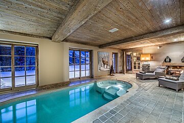 A luxurious indoor pool and jacuzzi room with rustic wood beams, stone floor, large windows to a snowy landscape, and a warm fireplace.