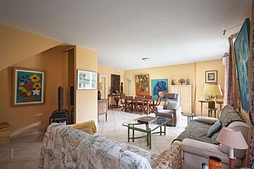 A warm, eclectic living and dining room with yellow walls, multiple artworks, patterned sofas, and a wooden dining set.