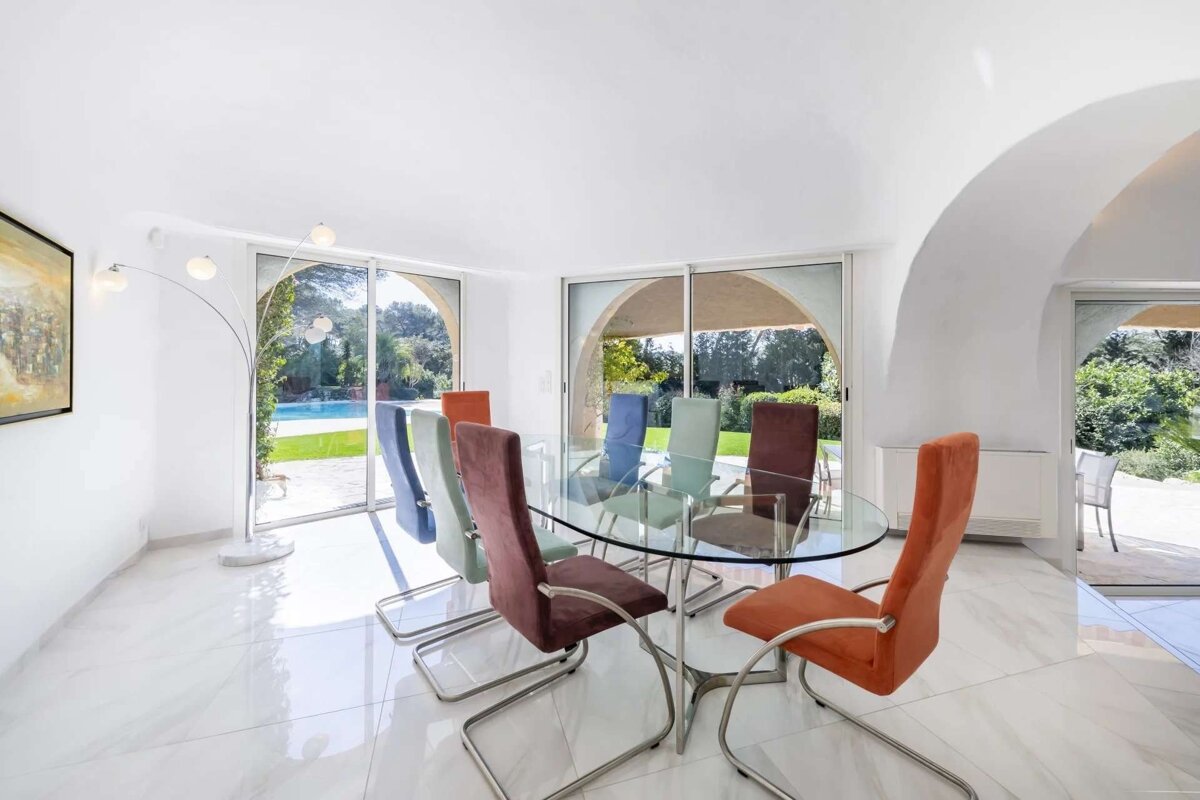 Bright, modern dining room with a glass table, colorful chairs, and arch-framed windows overlooking a sunny pool and garden.
