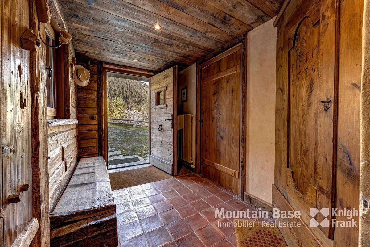 A rustic wooden cabin entryway with a bench, tiled floor, and an open door offering a glimpse of a lush green mountain landscape.
