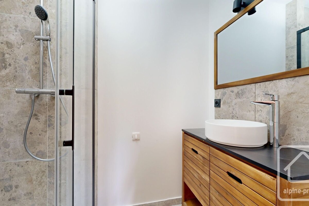 Modern bathroom with a glass shower, white walls, a wooden vanity with a white vessel sink, and a framed mirror above gray marble-look tiles.