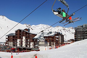 People on a ski lift ascend over a snowy mountain resort with chalet-style buildings and distant peaks under a clear blue sky.