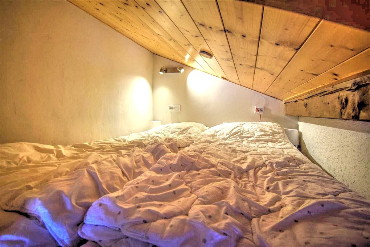 Cozy loft bed with an unmade white duvet under a low, sloped wooden ceiling.