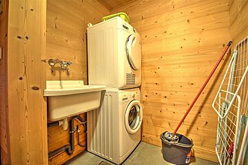 A rustic laundry room featuring wood paneling, a stacked washer and dryer, utility sink, mop and bucket, and a clothes drying rack.