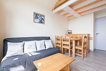 A bright living/dining room featuring a grey sofa, wooden coffee table, dining set, and sideboard. It has light walls and a decorative beamed ceiling.