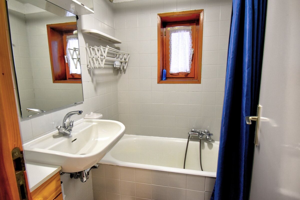 A tiled bathroom with a sink, bathtub, blue shower curtain, mirror, small window, and a wall-mounted drying rack.