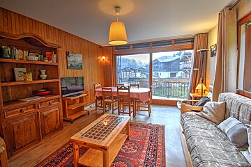 A cozy, wood-paneled living room and dining area featuring a patterned rug, TV, and a large balcony overlooking a snowy mountain village.